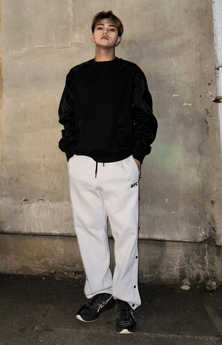 Jahan wearing a black sweatshirt and white sweatpands, standing with his hands in his pockets and looking down at the camera