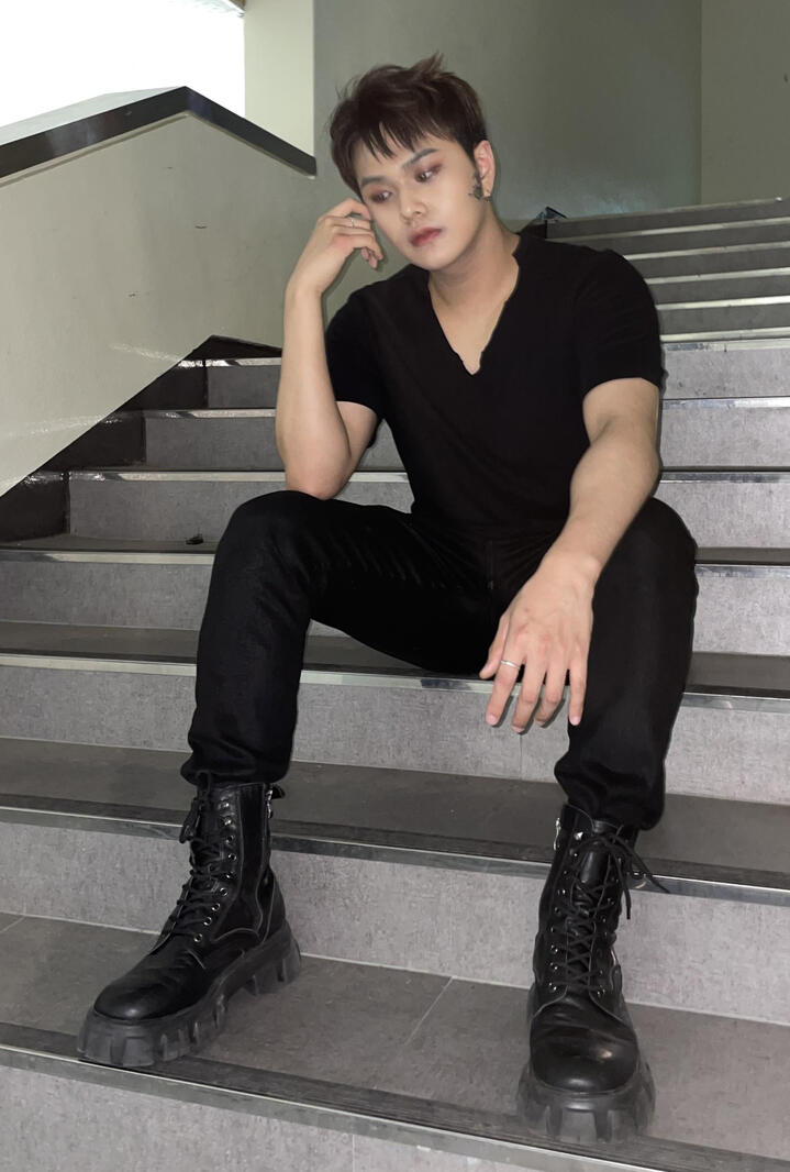 Jahan sitting on stairs, wearing a black v-neck tshirt and black pants, resting his elbow on his thigh and head on his hand