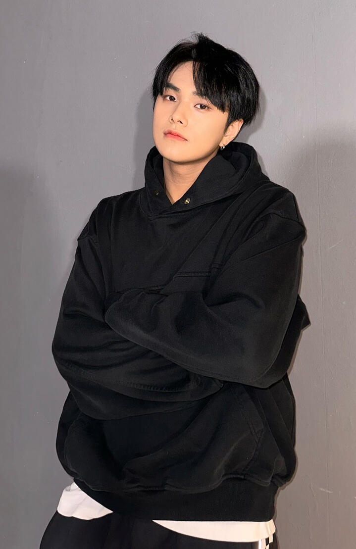 Jahan wearing a black hoodie and crossing his arms, leaning against a brick wall and looking into the camera