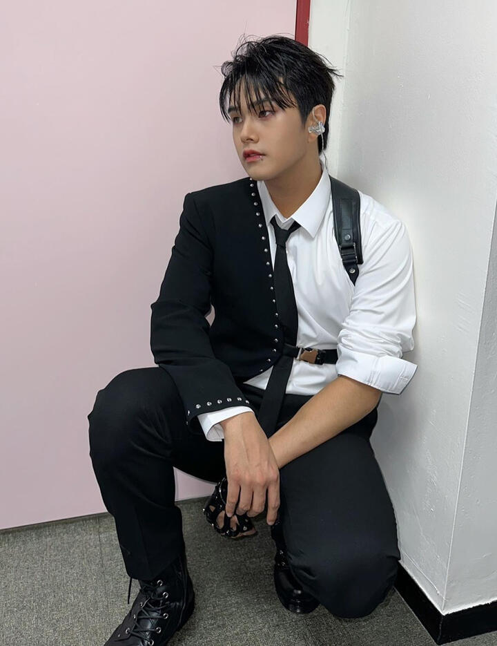 Jahan wearing a studded black half-jacket over a white button up and tie and black pants, knelt down and gazing to the side
