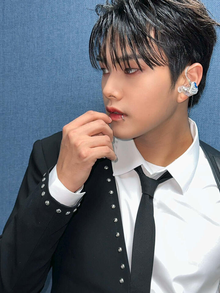 Jahan wearing a studded black half-jacket over a white button up and black tie with a lip ring, holding his hand loosely by his mouth and gazing to the side